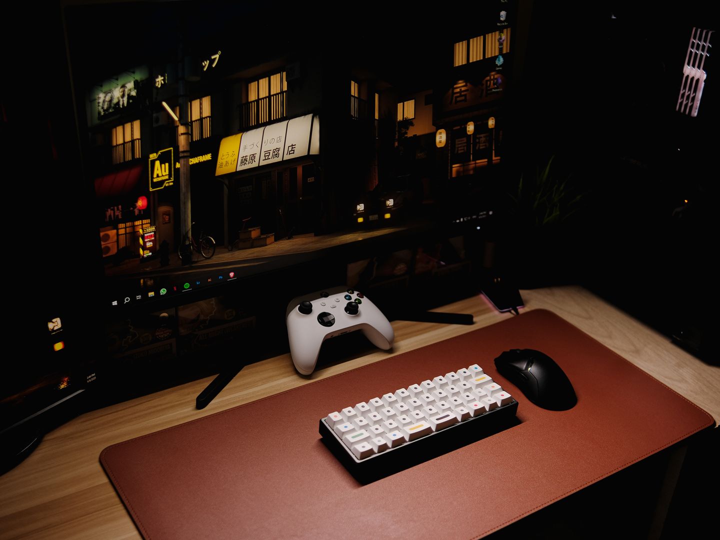 Close up of a gaming desk setup featuring a keyboard, controller, and monitor at night.
